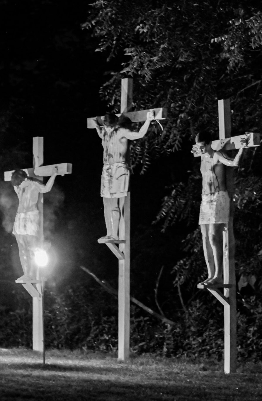 three men on crosses representing what it may have looked like to when Jesus was on the cross next to two thieves as they were crucified. The night is dark, torches illuminate the area. The men are covered only by loin coverings. They look anquished.